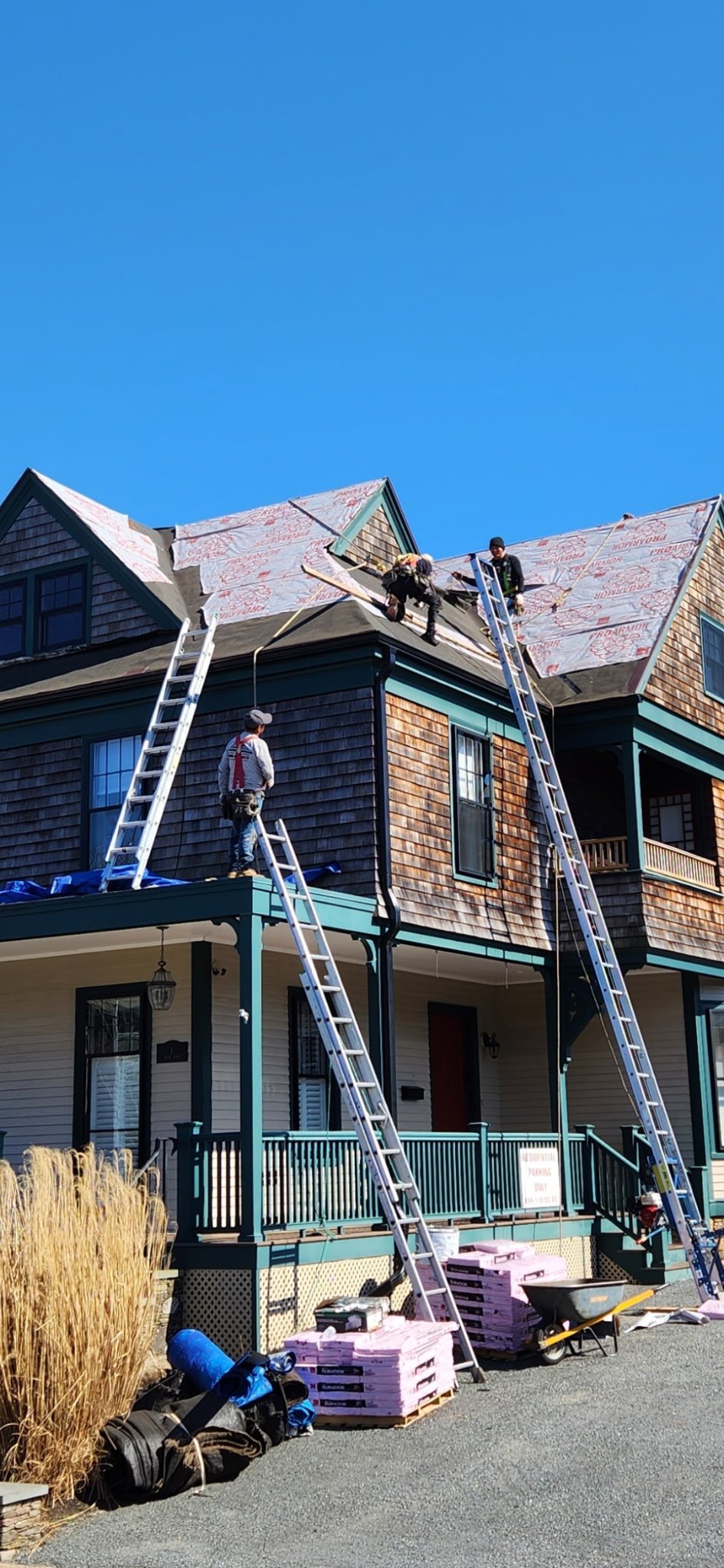 Workers on ladders installing roofing materials on a two-story house with wooden siding under clear blue sky
