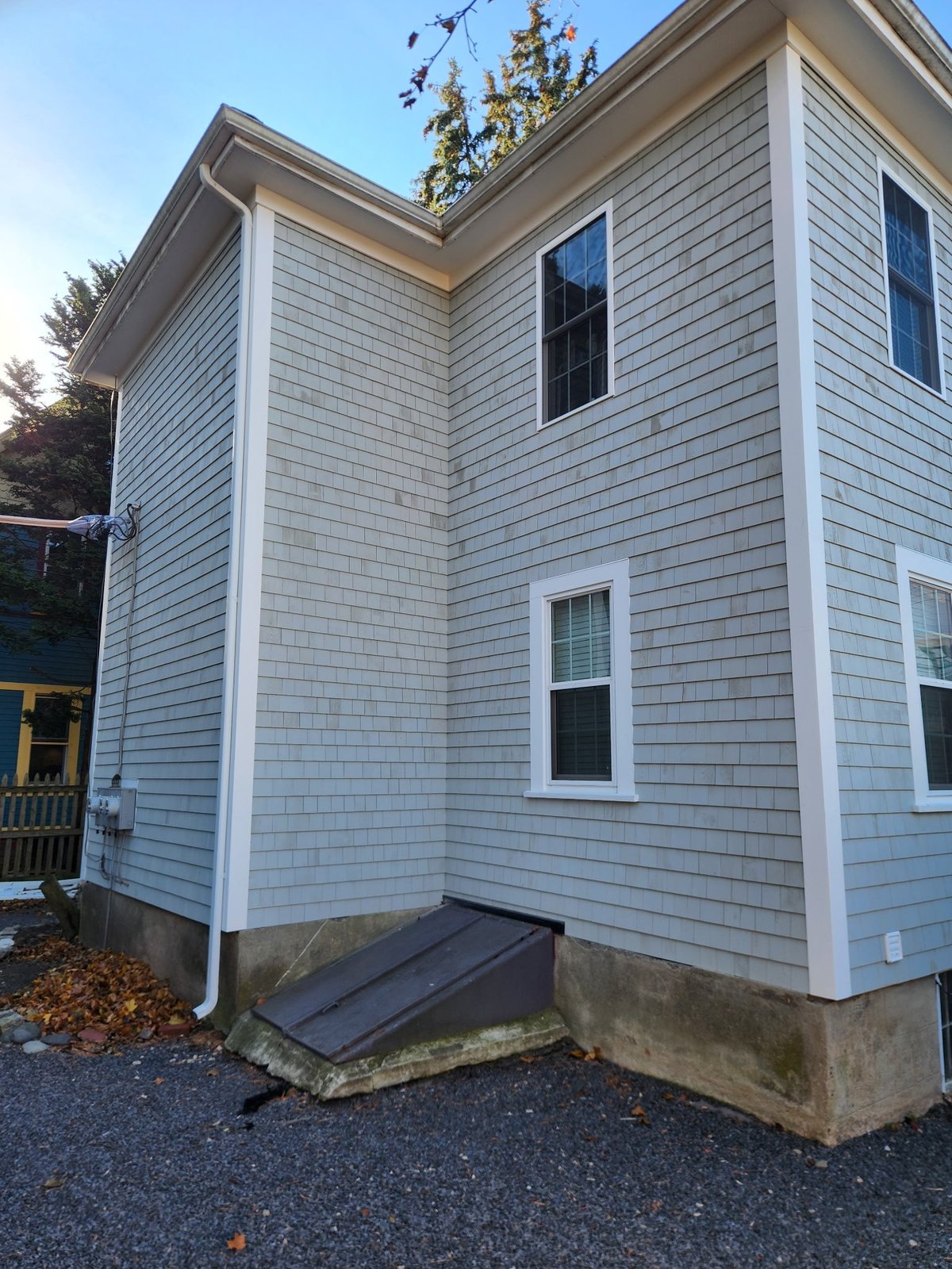 Two-story house with light blue-gray shingle siding, white trim, and dark windows against a clear sky.