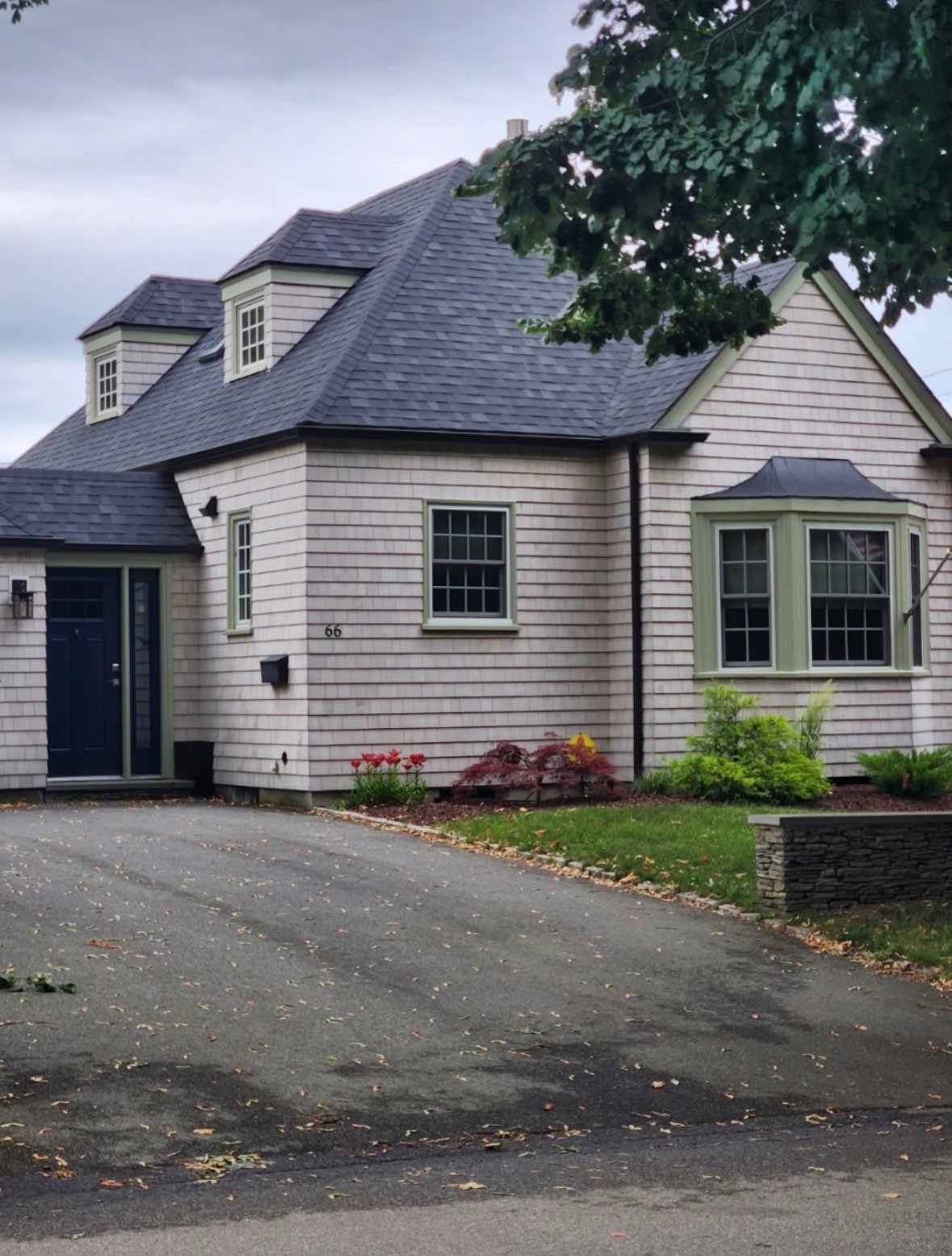 Modern farmhouse with white shingle exterior, dark roof, dormers, green shutters, and landscaping beside a driveway.